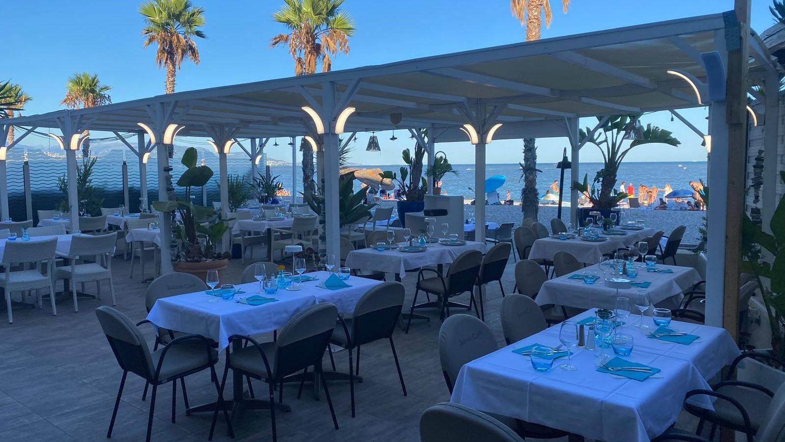 La terrasse de L&rsquo;Indigo dressée sous la pergola blanche, palmiers et plage de galets face à la Méditerranée
